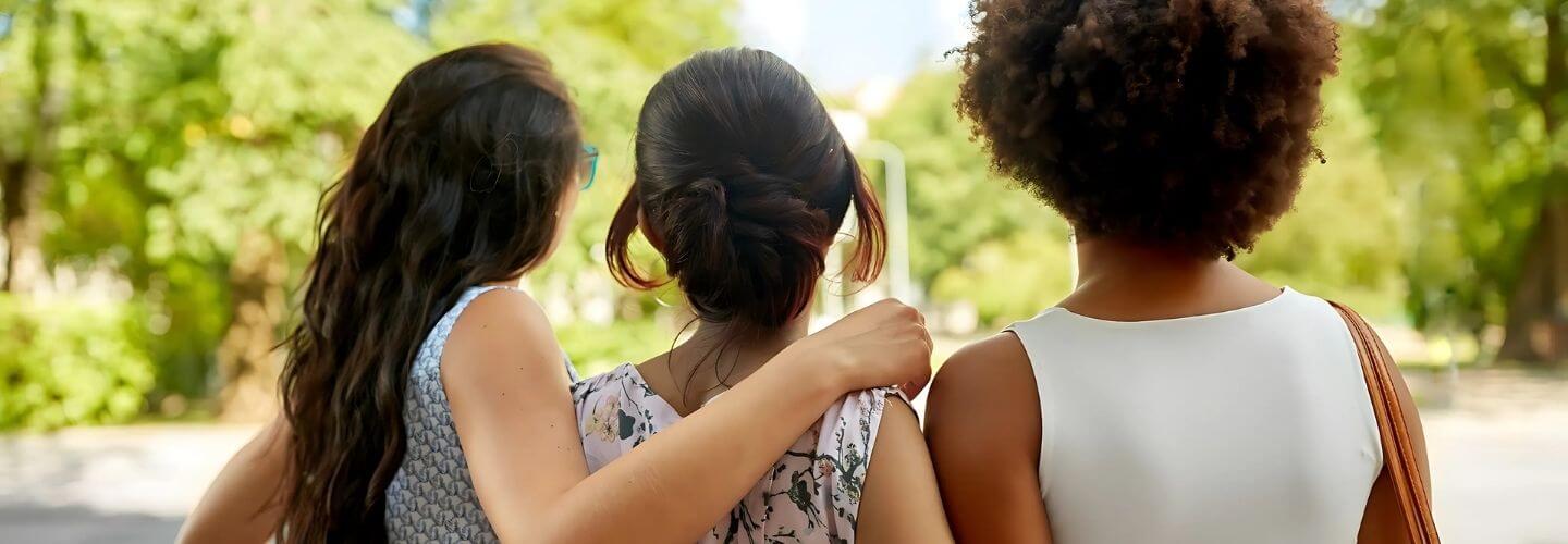 Three women standing side by side outdoors, one placing a supportive arm around another—symbolizing female friendship, unity, and the subtle complexities within sisterhood.