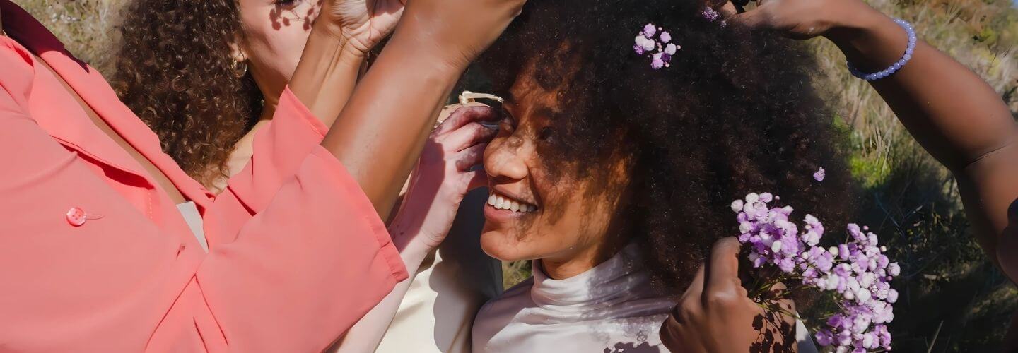 Smiling Black woman surrounded by friends gently placing flowers in her natural hair, symbolizing joy, community, and soft feminine care.
