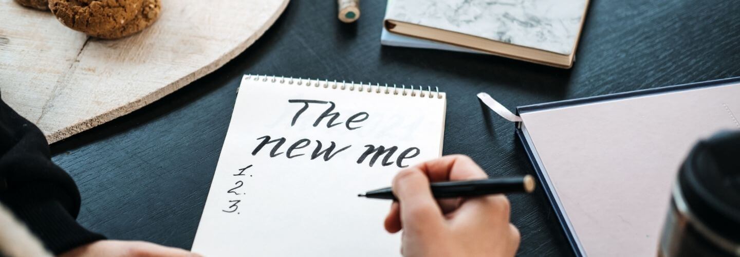 Person writing “The new me” on a spiral notepad with a pen, leaving numbered lines for a list, on a dark desk with books and cookies nearby.