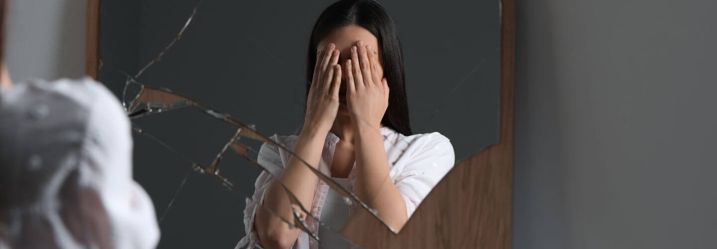 Woman covering her eyes in front of a broken mirror representing emotional unavailability to yourself and self-disconnection.